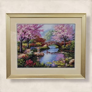 Puede incluir: Obra de arte en punto de cruz enmarcada que representa una serena escena de jardín japonés. La obra presenta un puente de madera sobre un estanque reflectante, rodeado de vibrantes cerezos en flor rosas y exuberante vegetación. El marco es dorado.