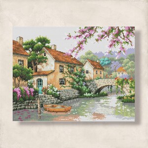 Puede incluir: Paisaje de punto de cruz que representa una escena de pueblo. La imagen muestra casas con tejados naranjas, un puente de piedra y un pequeño bote en el agua. La exuberante vegetación y los árboles de flores rosas contribuyen al idílico entorno.