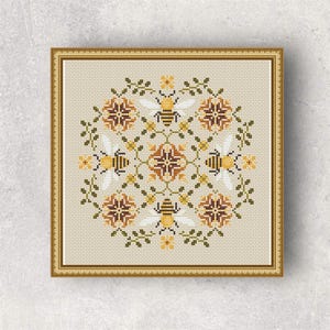 May include: Cross-stitch artwork with a symmetrical design of bees, flowers, and geometric shapes in yellow, brown, and green. The artwork is framed in a gold-coloured frame. The background is a light beige colour.