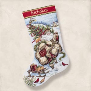 May include: Christmas stocking with a winter scene featuring Santa Claus carrying a sack. The stocking has the name "Nicholas" at the top. Presents on a sled and red birds are at the bottom. The background shows a snowy landscape.