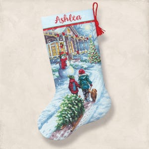 Könnte beinhalten: Hellblaue Weihnachtsstrumpf mit einer Winterszene mit einem Haus, einem Schneemann, Kindern und einem Hund. Das Wort "Ashlea" ist rot gestickt. Oben hängt eine rote Quaste.