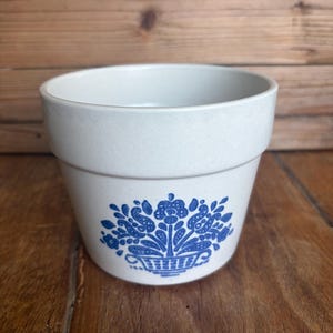 May include: A light beige ceramic planter with a wide, flared rim and a tapered body. The front of the planter features a blue printed graphic of a floral arrangement contained within a woven basket, showcasing intricate petal and leaf details.