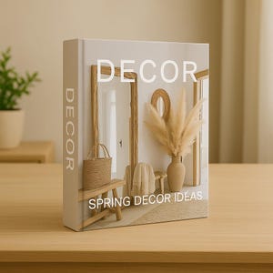 May include: A book titled "DECOR" with "SPRING DECOR IDEAS" printed on the cover. The cover features a miniature interior design scene with a mirror, bench, and vase with pampas grass. The book is beige and sits on a wooden table.