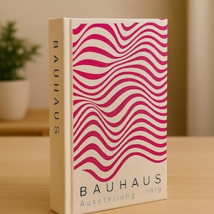 Könnte beinhalten: Ein Hardcover-Buch mit cremefarbenem Einband und einem wellenförmigen rosa Muster. Der Titel "BAUHAUS" ist vertikal auf dem Buchrücken und horizontal auf dem Cover zusammen mit "Ausstellung 1919" aufgedruckt. Das Buch liegt auf einer Holzoberfläche.