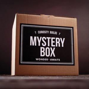 Puede incluir: Una caja misteriosa de cartón marrón con una etiqueta en blanco y negro. La etiqueta dice "MYSTERY BOX" en letras blancas grandes, con "Curiosity Builds" y "Wonder Awaits" arriba y abajo. La caja está sobre una superficie oscura.