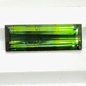 May include: A rectangular, faceted green tourmaline gemstone. The gem has a deep, rich green color with visible internal structures. The stone is set against a white background, with green foliage visible in the background.