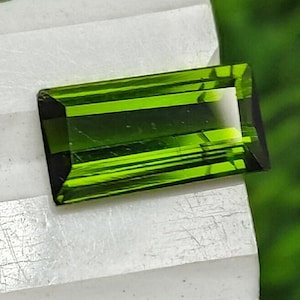 May include: A rectangular, emerald-green gemstone with a polished surface. The gem has a dark green hue and is set against a white surface with a blurred green background. The gemstone's facets reflect light, creating a brilliant appearance.