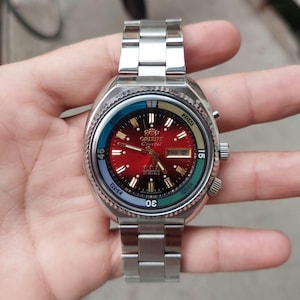 以下が含まれることがあります： 赤い文字盤と日付表示を備えた、ステンレススチール製のオリエントクリスタル自動腕時計。時計は、青、緑、黄色のマーキングが付いた回転ベゼルを備えています。時計バンドはステンレススチール製です。