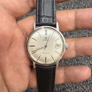 Pode incluir: Um relógio Omega Genève prateado com uma pulseira de couro preta. O mostrador do relógio é prateado com marcadores de hora e ponteiros pretos. A data é exibida às 3 horas. O relógio é segurado por uma mão.