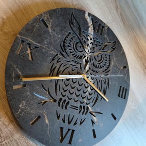 Puede incluir: Un reloj de pared redondo con una esfera gris oscuro con efecto mármol. El reloj presenta un diseño de búho recortado, números romanos y manecillas doradas. El diámetro del reloj es de aproximadamente 30 cm.