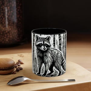 May include: Black and white ceramic coffee mug featuring a raccoon illustration. The raccoon is depicted in a detailed, linocut style, set against a backdrop of stylized trees. The mug has a black interior and is placed on a wooden surface.