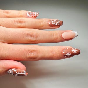 May include: A hand with festive, decorated fingernails. The nails feature a brown base with white gingerbread-themed designs, including houses, snowflakes, and a gingerbread man. One nail has a classic French manicure design.