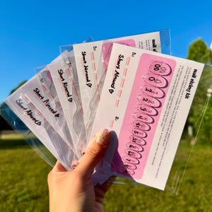 May include: A hand holding several clear plastic nail sizing kits. Each kit contains a pink card with clear nail tip sizes and the text "nail sizing kit". The cards are labeled "Short Almond".