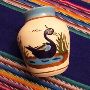 Puede incluir: Un jarrón de cerámica blanca con un borde azul y un diseño pintado de un pájaro azul en un estanque con juncos verdes. El jarrón está sobre una tela de rayas de colores.