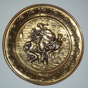 Può includere: Un piatto rotondo in metallo dorato con un design in rilievo di quattro figure. Le figure sono in una scena di un musicista che suona un liuto, con due donne e un uomo che ascoltano. Il piatto ha un bordo decorativo con un motivo ripetitivo.