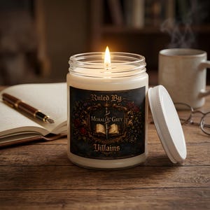 Op de afbeelding: Een brandende kaars in een glazen pot met een wit deksel. Het etiket toont een donker, versierd wapenschild met een zwaard, een open boek en rode rozen, met de tekst "Ruled By Morally Grey Villains" in een decoratief gouden lettertype.