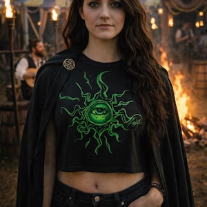 May include: A black crop top with a green eye design, paired with black jeans and a black cape. The eye design is surrounded by swirling lines. A gold brooch secures the cape. The background includes a bonfire and a festive setting.