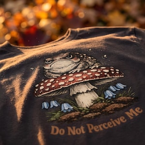 May include: A dark gray t-shirt featuring a graphic illustration of a grumpy toad sitting on a red and white spotted mushroom. Below the mushroom are small blue flowers and the text "Do Not Perceive Me" printed in a light tan, serif font.