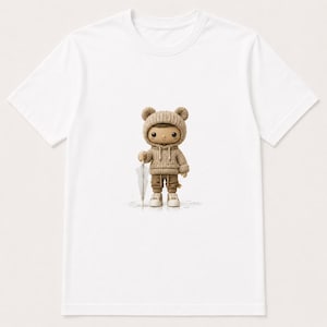 May include: A white t-shirt featuring a graphic of a small, stylized character wearing a beige knit hoodie with bear ears, matching pants, and light-colored sneakers. The character is holding a translucent umbrella and standing on a light surface.