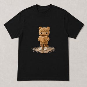 May include: A black short-sleeved t-shirt featuring a graphic of a small, tan, bear-eared character wearing a knit sweater and pants. The character holds an open book with the text "I AM NOT A ROBOT" printed on the pages, standing on a tan puddle.