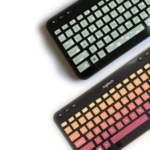 May include: Two black Logitech keyboards. The top keyboard features light green keys with small white daisy patterns. The bottom keyboard displays a gradient of orange, yellow, and pink keys. Both keyboards have standard QWERTY layouts.