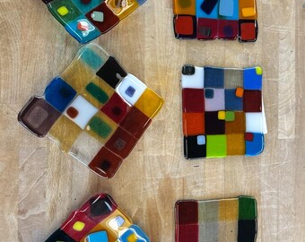 Könnte beinhalten: Sechs bunte Glasuntersetzer mit geometrischen Designs. Jeder Untersetzer hat eine einzigartige Anordnung von Quadraten in verschiedenen Farben, darunter Rot, Blau, Gelb und Grün. Die Untersetzer sind auf einer hellen Holzoberfläche angeordnet.