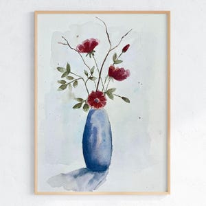 May include: A watercolor painting in a light wood frame depicting a blue vase containing three dark red flowers with green leaves and thin brown stems. The background is a soft, pale blue wash with subtle paint splatters and a cast shadow.