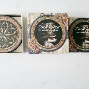 May include: A set of oracle cards and coasters. The oracle card boxes feature a steampunk design with a meerkat wearing a hat, the text "Zoologica The Steampunk Oracle", and the author's name. The coasters have a similar design.