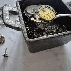 May include: A dark grey, square container filled with various small metal charms and a spoon. The spoon holds a gold-coloured clock charm. Other charms include gears, Eiffel Tower, and heart shapes. The container has two handles.
