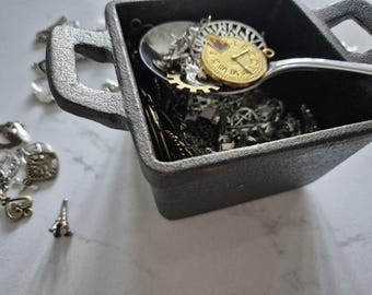 May include: A dark gray, square container filled with various small metal charms and a spoon. The spoon holds a gold-colored clock charm. Other charms include gears, Eiffel Tower, and heart shapes. The container has two handles.