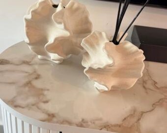 May include: Two off-white, abstract ceramic vases with a textured, organic design sit on a marble-patterned table. One vase holds black reeds. The table is part of a white cabinet with vertical lines.