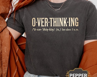 May include: Dark gray t-shirt with the words "O-VER-THINK-ING" in gold lettering. Below the text is the phonetic pronunciation and "See also: 3 a.m." The shirt has a vintage look and feel.