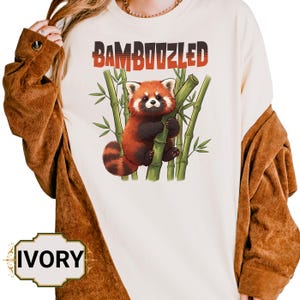 May include: Ivory sweatshirt with a red panda illustration clutching bamboo. The word "BAMBOOZLED" is printed above in red. The model is wearing a brown jacket. The word "IVORY" is printed on the bottom.