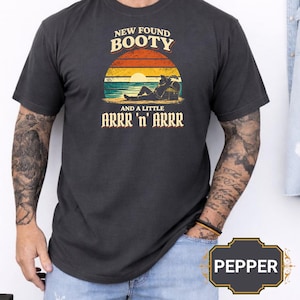 May include: Dark gray t-shirt with the text "NEW FOUND BOOTY" above a beach scene with a pirate and treasure chest. Below the scene is the text "AND A LITTLE ARRR 'n' ARRR". The shirt has short sleeves.
