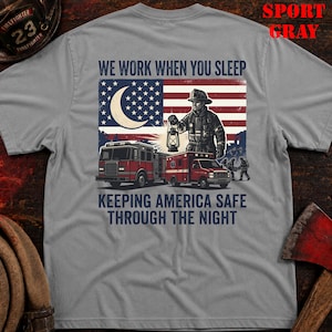 May include: Sport gray t-shirt featuring a graphic of a firefighter holding a lantern in front of an American flag, fire trucks, and the text "WE WORK WHEN YOU SLEEP KEEPING AMERICA SAFE THROUGH THE NIGHT."