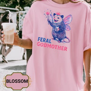May include: A light pink t-shirt featuring a whimsical illustration of an opossum with fairy wings, a crown, and a magic wand. The text "FERAL GODMOTHER" is printed below the opossum in blue and pink. The shirt is a casual wear item.