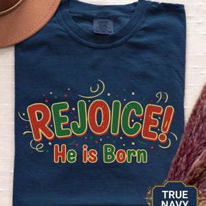 May include: A navy blue t-shirt featuring the text "REJOICE! He is Born" in red and green. The shirt has a crew neck and short sleeves. The fabric appears soft and comfortable. The t-shirt is a standard fit.