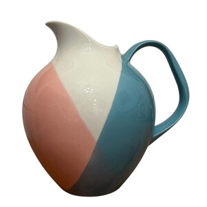 Puede incluir: Una jarra de cerámica con cuerpo redondeado y acabado brillante. La superficie está dividida en tres secciones diagonales de color: un rosa pastel suave a la izquierda, un panel central blanco y un azul turquesa vibrante en el lado derecho.