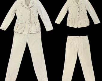 May include: A beige two-piece suit, including a jacket and pants, displayed on a black background. The jacket has a button-up front, multiple pockets, and long sleeves. The pants are straight-legged.