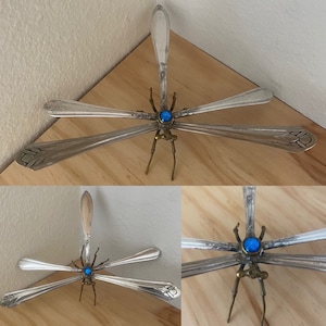 May include: A dragonfly sculpture made from recycled silverware with a blue gemstone accent. The dragonfly is posed with its wings spread out.