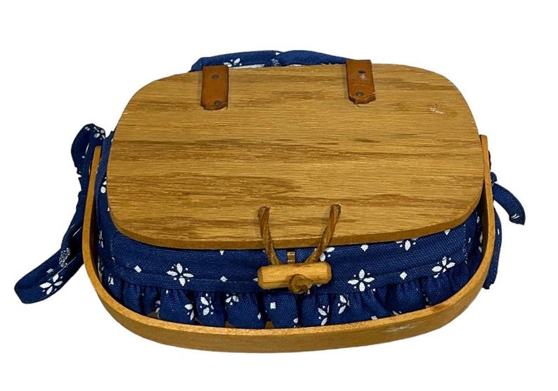 May include: A blue and white fabric-lined wooden basket with a hinged lid and a wooden handle. The basket has a white floral pattern on the fabric lining.