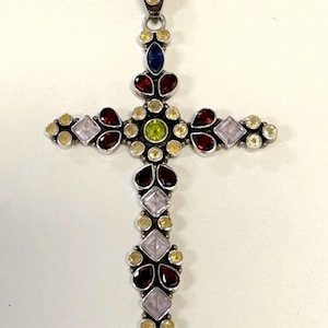 May include: Silver-toned pendant in the shape of a cross, adorned with various gemstones. The gemstones include red, yellow, green, pink, and blue stones. The pendant has a loop at the top for a chain.