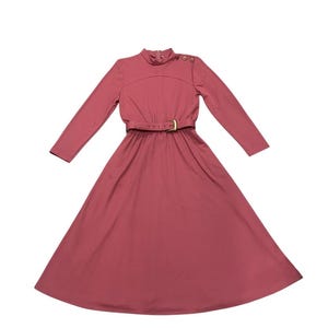 May include: A dusty rose-colored dress with long sleeves and a high neckline. The dress features a fitted bodice with a belt and a flared skirt. Gold-colored buttons adorn the shoulders. The dress is made of a soft, flowing fabric.