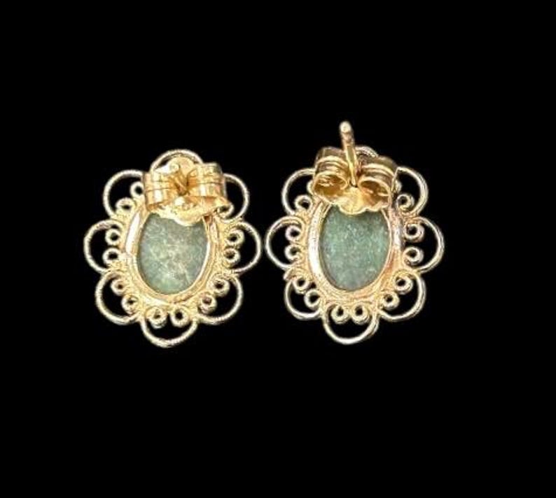 May include: A pair of gold-colored stud earrings. Each earring features an oval, light green stone set within a decorative, openwork frame. The earrings have a post and butterfly backing.
