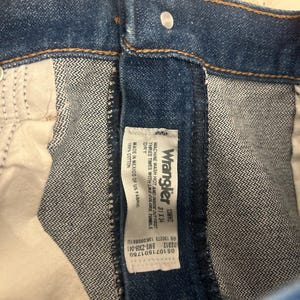 Op de afbeelding: Close-up van een blauwe spijkerbroek met een zichtbaar Wrangler-label. Het label geeft details over de jeans, inclusief de maat 31x34 en wasinstructies. De jeans is gemaakt van 100% katoen.