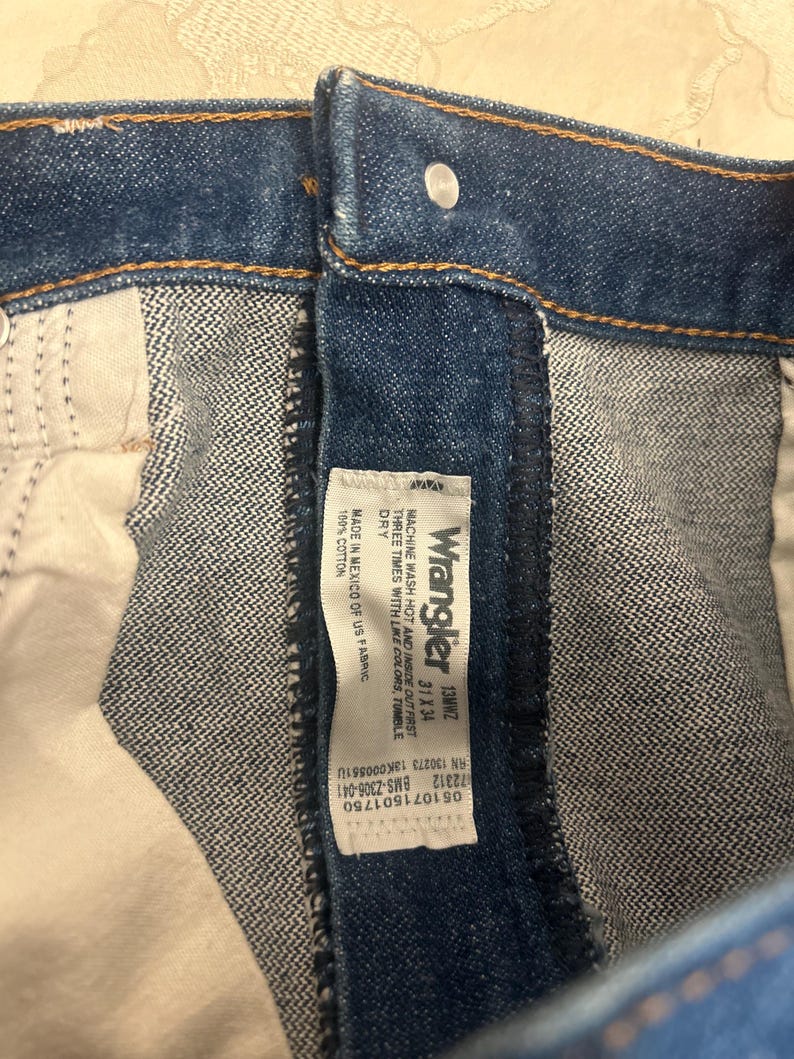 Op de afbeelding: Close-up van een blauwe spijkerbroek met een zichtbaar Wrangler-label. Het label geeft details over de jeans, inclusief de maat 31x34 en wasinstructies. De jeans is gemaakt van 100% katoen.