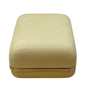 May include: A rectangular, off-white jewelry box with a textured surface. The box has a gold-colored metal trim around the base. The box is closed and appears to be empty.