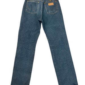 Op de afbeelding: Donkerblauwe denim jeans met een klassiek ontwerp met vijf zakken. De jeans hebben een bruine leren patch en het Wrangler-logo. De achterzakken hebben een W-vormige stiksels. De jeans liggen plat op een witte achtergrond.