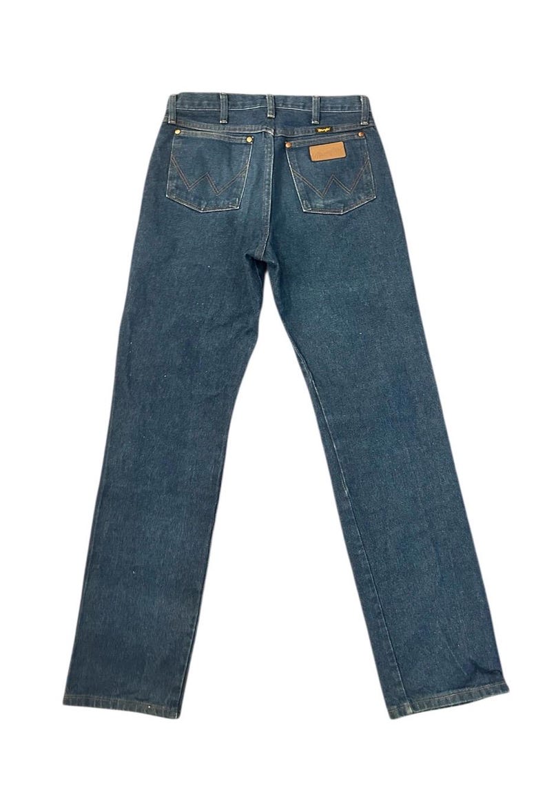 Op de afbeelding: Donkerblauwe denim jeans met een klassiek ontwerp met vijf zakken. De jeans hebben een bruine leren patch en het Wrangler-logo. De achterzakken hebben een W-vormige stiksels. De jeans liggen plat op een witte achtergrond.