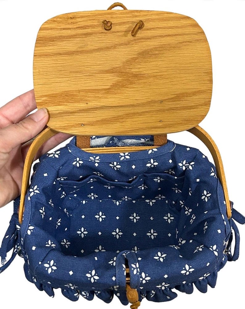 May include: A blue and white fabric-lined basket with a wooden lid and handles. The basket has a floral pattern and is perfect for storage or carrying small items.